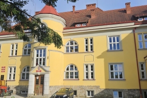 Żółty budynek z czerwonym dachem, wieżyczką i dużymi oknami. Przed wejściem widać ciągnik z przyczepą oraz samochód, a wokół budynku znajdują się drzewa.