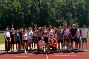 Grupa młodzieży pozuje do wspólnego zdjęcia na bieżni lekkoatletycznej. Część osób trzyma medale i nagrody, a w tle widać drzewa oraz słoneczne niebo.