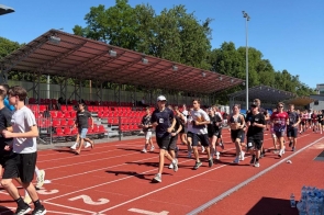 Grupa młodzieży biegnie po czerwonej bieżni lekkoatletycznej na stadionie. W tle widać trybuny z czerwonymi krzesełkami, drzewa oraz słoneczne niebo.
