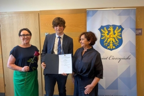 Trzy osoby pozują do zdjęcia. Młody mężczyzna trzyma dyplom, a za nim widoczny jest baner z herbem powiatu.