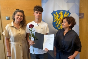 Trzy osoby pozują do zdjęcia. Młody mężczyzna trzyma różę i dyplom, a za nim widoczny jest baner z herbem powiatu.