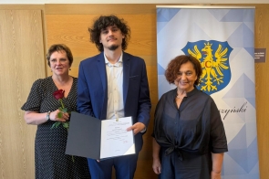 Trzy osoby pozują do zdjęcia. Młody mężczyzna trzyma dyplom, a za nim widoczny jest baner z herbem powiatu.