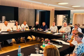 Osoby siedzą przy stołach w sali konferencyjnej. Na stołach widać dokumenty, laptopy, wodę, filiżanki oraz drobne dekoracje.
