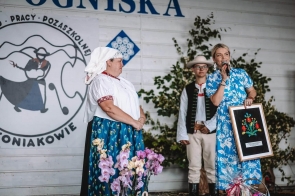 Aneta Legierska-Bujok Przewodnicząca Komisji Komisja Kultury, Sportu i Promocji  Powiatu przemawia do mikrofonu i trzyma pamiątkowy obraz,  Ewa Macoszek-Dragon Dyrektor Ogniska Pracy Pozaszkolnej w Koniakowie. W tle widoczna jest dekoracja nawiązująca do tradycji oraz jubileuszu 50-lecia.