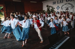 Dzieci i młodzież w strojach regionalnych tańczą na scenie podczas uroczystości. W tle widać muzyków oraz dużą dekorację z napisem nawiązującym do 50-lecia ośrodka, a na pierwszym planie stoi instrument klawiszowy.