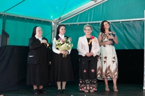 Na scenie stoją cztery kobiety podczas uroczystości. Dwie z nich są w strojach zakonnych, jedna trzyma bukiet kwiatów, a pozostałe osoby mają przypięte okolicznościowe wstążki. W tle widać turkusowe zadaszenie namiotu.