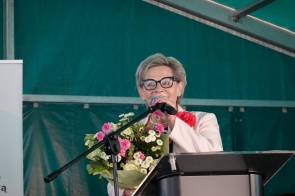 Beata Macura Członkini Zarządu przemawia do mikrofonu podczas uroczystości pod namiotem. Trzyma bukiet kwiatów.