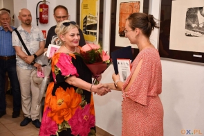 Dwie kobiety stoją w pomieszczeniu i podają sobie ręce. Jedna trzyma kwiaty, w tle widać inne osoby.