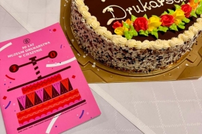 Okrągły tort z kolorową dekoracją na stole, obok różowa kartka z napisem.