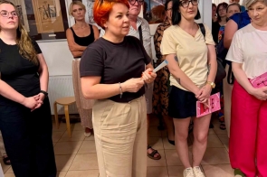 Grupa osób stoi w pomieszczeniu. Na środku kobieta przemawia, obok stoją inne osoby. W tle widać ściany i obrazy.