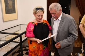 Kobieta i mężczyzna stoją w pomieszczeniu. Oboje trzymają kartkę papieru patrząc na nią. 