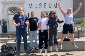 Pięć osób uchwycono podczas skoku przed budynkiem muzeum. W tle widoczna jest jasna elewacja z dużym napisem „MUSEUM”, schody oraz elementy małej architektury.
