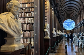 Wnętrze zabytkowej biblioteki z wysokimi regałami pełnymi książek. Wzdłuż sali ustawione są popiersia, a w głębi widoczna jest podświetlona kula ziemska.
