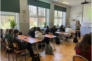 Uczniowie siedzą przy ławkach w sali lekcyjnej. Z przodu stoi osoba prowadząca, obok ekranu z wyświetloną prezentacją.