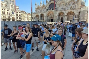 Grupa młodzieży stoi na placu przed bazyliką. W tle widać bogato zdobioną fasadę bazyliki, wysokie maszty oraz tłum turystów pod słonecznym, błękitnym niebem.