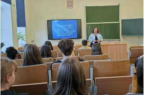 Uczniowie siedzą w sali wykładowej. Z przodu stoi osoba prowadząca, obok ekranu z wyświetloną prezentacją i tablic szkolnych.
