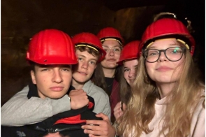 Grupa młodych osób ubranych w czerwone kaski ochronne, pozujących do wspólnego zdjęcia we wnętrzu o ciemnych ścianach. Uczestnicy stoją blisko siebie i patrzą w obiektyw aparatu.