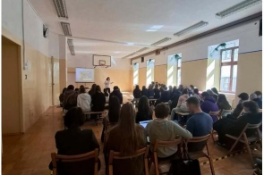 Grupa osób siedzi na krzesłach w sali gimnastycznej. Z przodu sali stoi osoba przy ekranie z wyświetloną prezentacją.