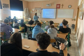 Uczniowie siedzą w ławkach w sali lekcyjnej. Z przodu stoi osoba prowadząca, obok ekranu z wyświetloną prezentacją.