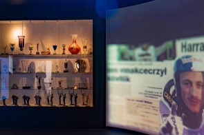 Wnętrze wystawy poświęconej skokom narciarskim: po lewej podświetlona gablota z pucharami i statuetkami, a po prawej projekcja filmu ze skoczkiem narciarskim.