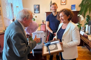 Kilka osób znajduje się w gabinetowym wnętrzu. Kobieta w białej marynarce trzyma upominek w ramce, starszy mężczyzna trzyma bukiet kwiatów, a obok stoi młody mężczyzna. W tle widoczne są drewniane meble, rośliny, godło i plakat na ścianie.