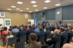 Sala konferencyjna z siedzącą publicznością. Przy mównicy stoi osoba w mundurze, a w tle widoczne są banery Policji i Powiatu Cieszyńskiego, flagi oraz godła.