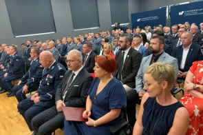W pierwszych rzędach widoczni są policjanci w mundurach oraz osoby w strojach formalnych. W tle znajdują się granatowe ścianki Uniwersytetu Śląskiego.