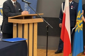 Mężczyzna stoi przy drewnianej mównicy w sali konferencyjnej. Obok znajduje się mikrofon, flaga Polski, flaga z herbem oraz osoba w mundurze. W tle widoczny jest niebieski baner i ciemne rolety.