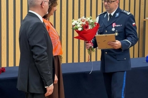 Trzy osoby stoją w sali uroczystości. Osoba w mundurze trzyma bukiet kwiatów i oprawiony dokument, a nad stołem widoczne jest godło Polski.