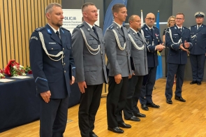 Policjanci w mundurach stoją w szeregu w sali konferencyjnej. Część osób trzyma teczki. W tle znajduje się baner Policji, flaga oraz stół z kwiatami.