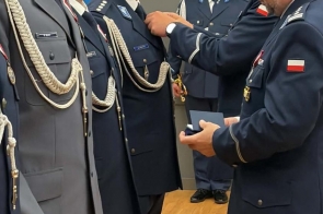 Policjanci w mundurach stoją w szeregu. Jedna osoba przypina odznaczenie do munduru, trzymając w dłoni otwarte etui. W tle widoczni są kolejni funkcjonariusze oraz szara ściana sali.