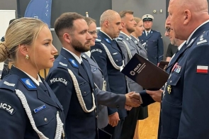 Policjanci w mundurach stoją w szeregu. Jedna osoba trzyma teczkę, a dwie osoby podają sobie ręce. W tle widoczne są kolejne osoby w mundurach oraz ciemne rolety.