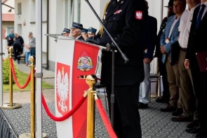 Osoba w mundurze stoi przy mównicy pod niebieskim namiotem. Przy mównicy znajduje się mikrofon, czerwono-biała dekoracja i godło, a w tle widoczne są inne osoby stojące na scenie.