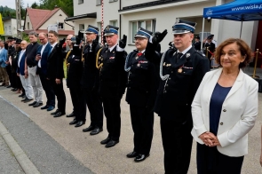 Grupa osób stoi w szeregu przed budynkiem podczas uroczystości. Część uczestników ma na sobie mundury i salutuje, a po prawej stronie stoi kobieta w białej marynarce.