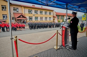 Osoba w mundurze stoi przy mównicy pod niebieskim namiotem. Przed budynkiem ustawiony jest szyk mundurowych, a po lewej stronie widoczna jest grupa osób w czerwonych strojach.