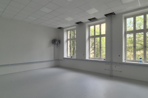 Jasne, wyremontowane pomieszczenie w jasnej kolorystyce, z dużymi oknami i nową podłogą. W suficie widoczne są częściowo otwarte panele, a przy ścianach znajdują się elementy instalacji.