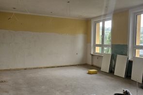 Pomieszczenie w trakcie remontu, z dużymi oknami i odsłoniętą instalacją pod sufitem. Przy ścianie ustawione są zdemontowane grzejniki, a na podłodze widoczne są materiały i elementy budowlane.