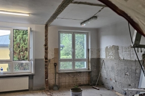 Pomieszczenie w trakcie remontu, z odsłoniętym fragmentem ściany i sufitu oraz częściowo skutymi płytkami. Widać duże okna, grzejnik oraz narzędzia budowlane