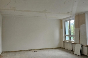 Pomieszczenie w trakcie remontu, z jasnymi ścianami, dużymi oknami i odsłoniętą instalacją pod sufitem. Przy ścianie widoczne są zdemontowane grzejniki, a podłoga nie jest wykończona.