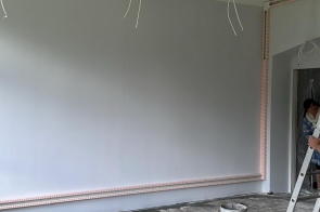 Pomieszczenie w trakcie remontu, z odsłoniętymi przewodami pod sufitem i niewykończoną podłogą. Przy ścianie widoczna jest drabina, wiadro oraz elementy budowlane.