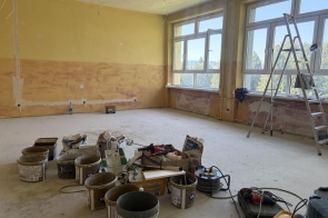 Pomieszczenie w trakcie remontu, z dużymi oknami, drabiną i odsłoniętymi przewodami pod sufitem. Na podłodze widoczne są wiadra, worki z materiałami budowlanymi, narzędzia oraz przedłużacze.