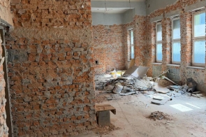 Pomieszczenie w trakcie remontu, z odsłoniętymi ceglanymi ścianami i przewodami zwisającymi z sufitu. Na podłodze widoczne są gruz, wiadra oraz materiały budowlane, a po prawej stronie znajdują się duże okna.