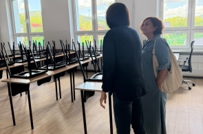 Jasna sala lekcyjna przygotowywana do nowego roku szkolnego. Krzesła ustawione są na ławkach, na podłodze widać jasne panele, a przy dużych oknach stoją dwie rozmawiające osoby.