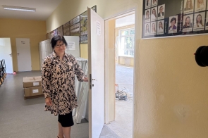 Kobieta stoi przy otwartych drzwiach prowadzących do remontowanej sali. W korytarzu widoczne są szafki, kartony i elementy wyposażenia, a w pomieszczeniu za drzwiami trwają prace przygotowawcze.