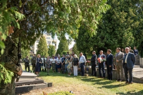 Uczestnicy uroczystości stoją na cmentarzu wśród zieleni. Przy grobach widoczni są przedstawiciele władz, żołnierze, harcerze oraz mieszkańcy, a jedna z osób przemawia przy mikrofonie.