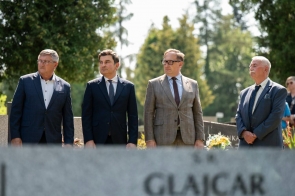 Czterech mężczyzn w garniturach stoi na cmentarzu podczas uroczystości. Na pierwszym planie rozmyta jest płyta nagrobna z napisem, a w tle widoczne są drzewa i kwiaty przy grobach.