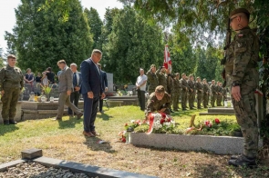 Mężczyzna w granatowym garniturze patrzy w kierunku grobu, przy którym żołnierz składa biało-czerwony wieniec. W tle stoją żołnierze ustawieni w szeregu oraz poczet sztandarowy.