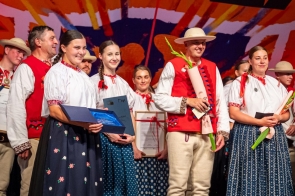 Grupa osób w strojach regionalnych stoi na scenie. Na pierwszym planie dwie kobiety trzymają otwarte teczki, mężczyzna w kapeluszu trzyma bukiet kwiatów, a stojąca obok kobieta – kwiat i kopertę.