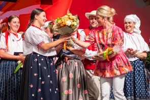 Kobieta w różowej bluzce w kwiaty przekazuje bukiet kobiecie w stroju regionalnym. Wokół stoją pozostałe osoby w strojach regionalnych, obserwujące wręczenie kwiatów.