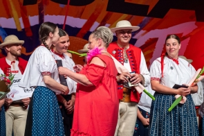 Kobieta w czerwonej sukience wręcza list gratulacyjny kobietom w strojach regionalnym. Wokół stoją pozostałe osoby w strojach regionalnych, część z kwiatami i dokumentami w dłoniach.